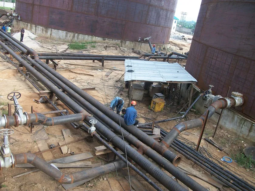 Large-bore mild steel water pipeline installation beside industrial storage tanks